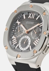 Orologio Guess con cassa in acciaio inossidabile argento, quadrante in nero testurizzato, dettagli in oro rosa, numeri romani sovradimensionati e cinturino in gomma.