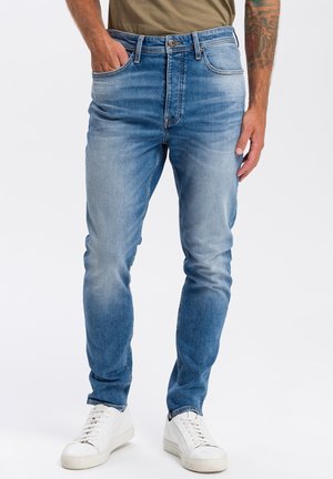 Man draagt lichtblauwe verwassen slim-fit jeans met een bruin shirt, witte sneakers en een zichtbaar getatoeëerde arm.