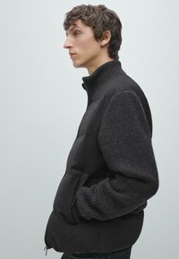 Young man in profile wearing a dark gray padded jacket with ribbed sleeves and collar, hands in pockets, against a plain light background.