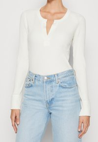 Vit ribbad långärmad topp med en henley-krage och en enkel framknapp, kombinerad med ljusblå högmidjade jeans.