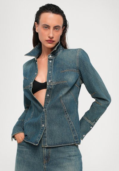 Dunkelblaue Jeansjacke mit hohem Kragen, Fronttaschen und silbernen Druckknöpfen. Leicht taillierte Passform mit sichtbaren Nähdetails.