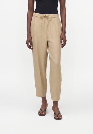 Vrouw met beige broek met trekkoord, zwart mouwloos topje en donkere sandalen met vierkante neus, staand tegen een effen witte achtergrond.