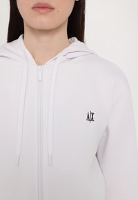 Biela mikina na zips vyrobená z mäkkej látky, s predným zipsom, bielymi šnúrkami a malým čiernym logom "A/X" na ľavej hrudi.