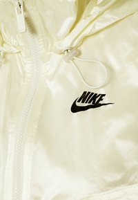 Ljusgul Nike-jacka med dragkedja framtill, justerbar snörning och svart logotyp på bröstet. Slät, glänsande tygtextur.