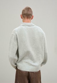Szürke kötött pulóver galléros nyakkal, bordás mandzsettákkal és hemésszel, valamint puha textúrával, barna bőszárú nadrággal viselve.
