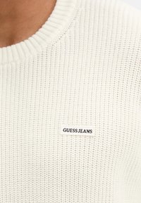 Maglione bianco lavorato a maglia con scollo a coste. Presenta una piccola etichetta nera con la scritta "GUESS JEANS" sul davanti. Design del tessuto strutturato.