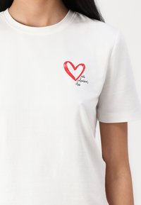 Balta medvilninė marškinė su raudonu širdies grafiku ir tekstu "antivalentinų klubas", atspausdintu juoda spalva šalia kairiojo krūtinės.