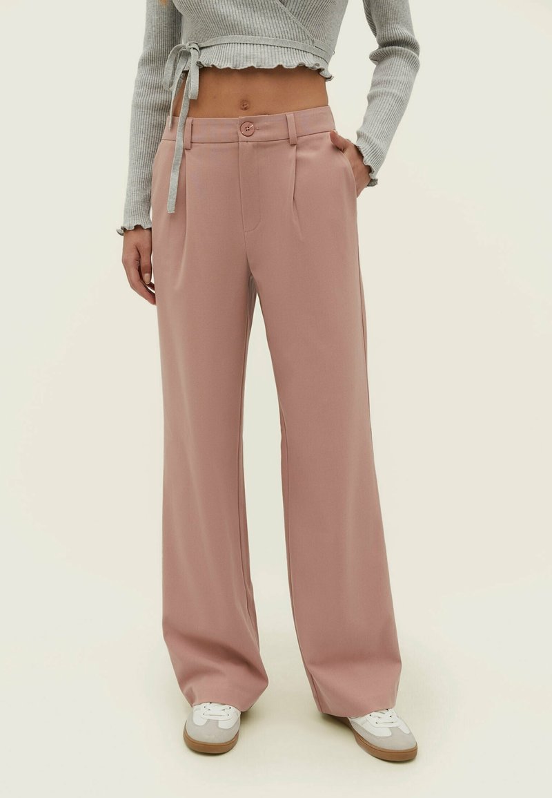 Femme portant un pantalon taille haute large rose poudré, un pull court gris cache-cœur et des baskets blanches avec semelles en gomme.