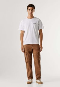 Wit katoenen T-shirt met korte mouwen en een kleine zwarte grafische print, gecombineerd met bruine cargobroek met zijzakken. Sneakers maken de outfit compleet.