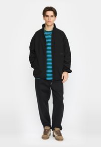 Zwarte jas met een hoge kraag en een borstzak, gedragen over een gestreept blauw en teal shirt, gecombineerd met zwarte broek en bruine sneakers.