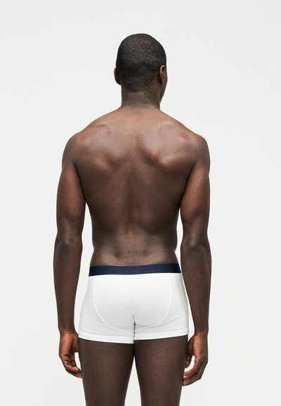 Μυώδης άνδρας που στέκεται πλάτη στην κάμερα, φορώντας λευκά ανδρικά εσώρουχα με σκουρόχρωμη μέση, μπροστά σε απλό φωτεινό φόντο.
