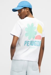 Γυναίκα που φοράει λευκό μπλουζάκι με πολύχρωμα λουλούδια και φύλλα και το κείμενο "Perugia", ανοιχτό μπλε καπέλο και ανοιχτόχρωμα τζιν σορτς.