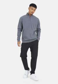 Felpa grigia con zip e maniche lunghe, pantaloni cargo neri con tasche con zip e sneakers bianche. Design casual e aderente.