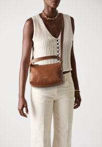 Femeie îmbrăcată cu un top fără mâneci, tricotat în model cable-knit de culoare crem, pantaloni de culoare albă cu nuanțe off-white, colier din lanț de aur și purtând o geantă crossbody din piele maro.