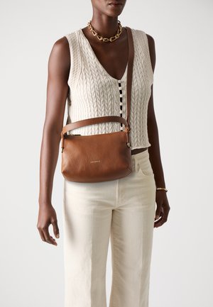 Woman wearing cream cable-knit sleeveless top, off-white pants, gold chain necklace, and carrying a brown leather crossbody bag.