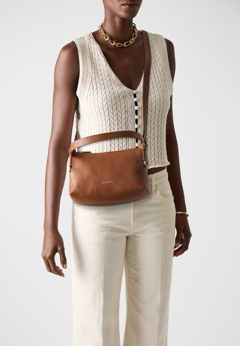 Femeie îmbrăcată cu un top fără mâneci, tricotat în model cable-knit de culoare crem, pantaloni de culoare albă cu nuanțe off-white, colier din lanț de aur și purtând o geantă crossbody din piele maro.