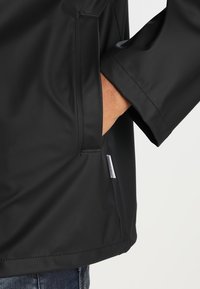 Svart vattentät jacka med en sidficka, slät yta, raglanärm-design och en liten varumärkesetikett vid nederkanten.