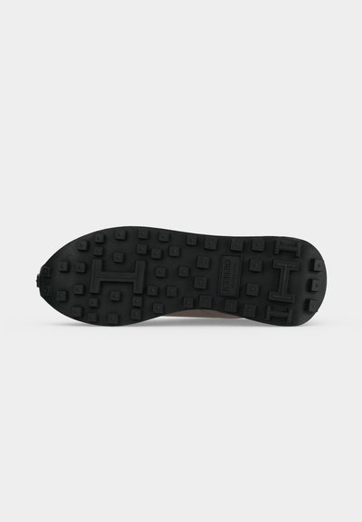 Semelle en caoutchouc noire d'une chaussure avec des crampons carrés et le nom de la marque embossé près du centre sur un fond clair uni.