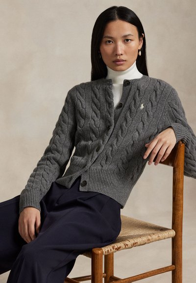 Nő ül egy fa széken, szürke fonott mintájú kardigánt, fehér garbót, tengerészkék nadrágot és arany karika fülbevalókat visel, semleges háttér.