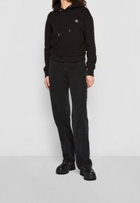 Sudadera negra con cordones y logo, combinada con jeans negros de pierna recta y calzado negro grueso. Tela suave, diseño casual.