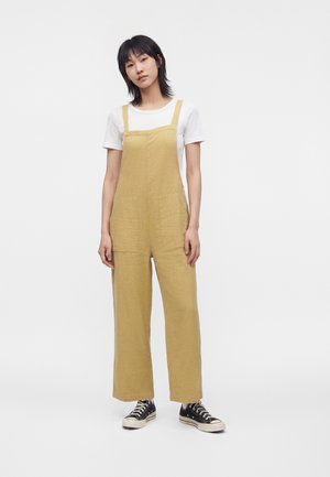 Tânără stând în picioare, purtând salopetă galben-muștar cu picior larg, tricou alb cu mânecă scurtă și adidași negri high-top pe fundal alb.