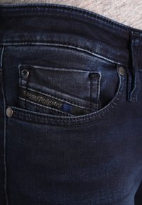 Dark blue denim jeans featuring a small front pocket with a black interior and blue label. The fabric has a slight sheen and smooth texture.