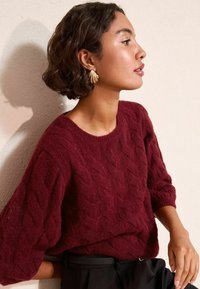 Burgundy kötött pulóver, hajtogatott mintával és széles ujjal, fekete nadrággal párosítva. Arany fülbevalók figyelemfelkeltő részletet adnak.
