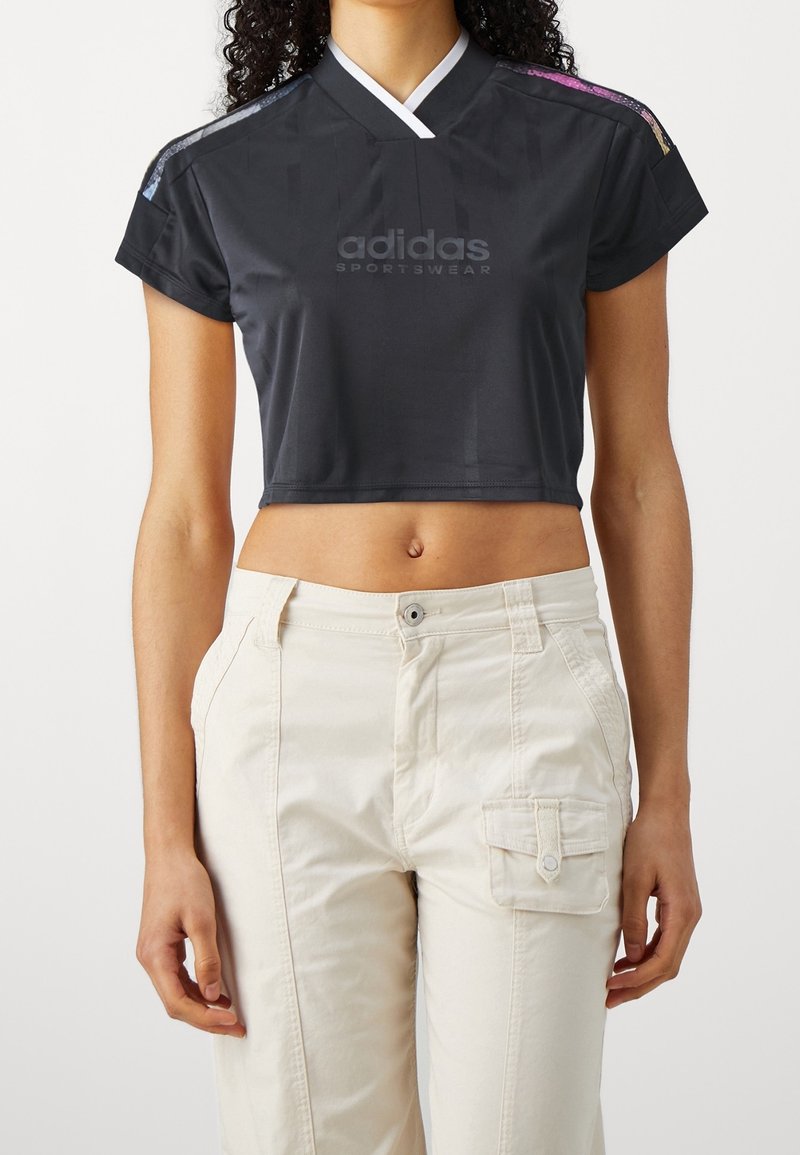 Černý crop top vyrobený z látky s výstřihem do V a krátkými rukávy. Na přední straně je logo Adidas; barevné akcenty na ramenou. V kombinaci s světle béžovými kalhotami.