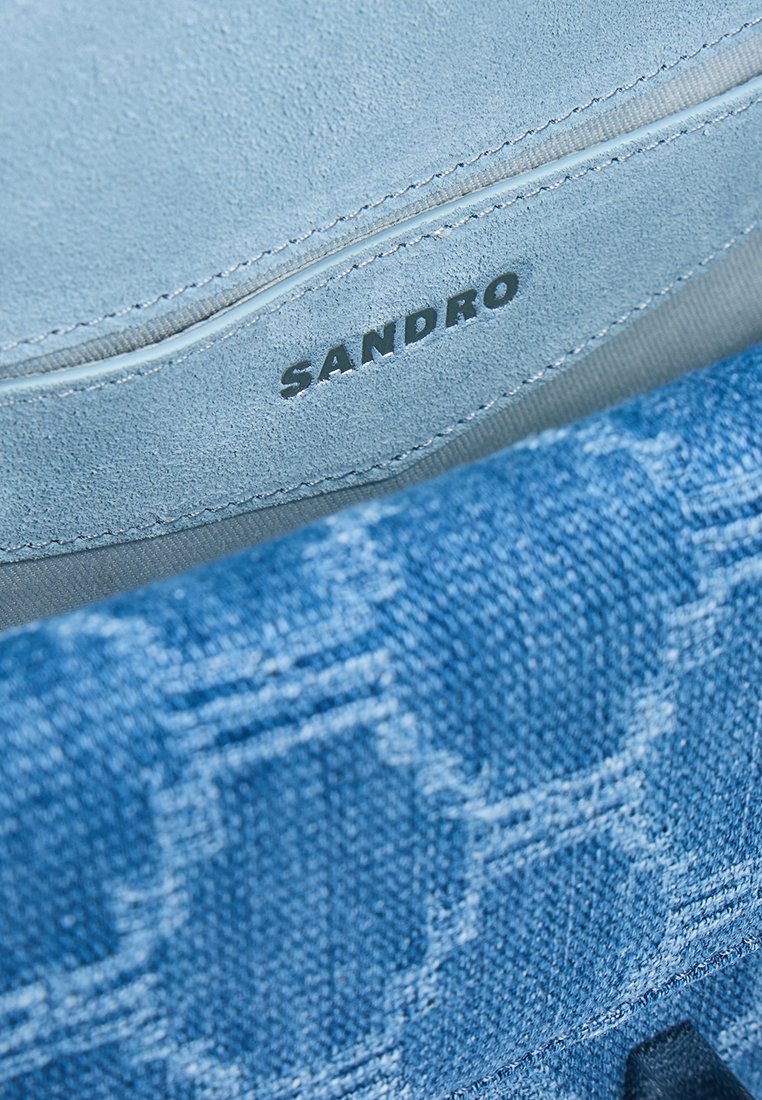 Blue denim bag interior featuring a smooth, light blue suede lining. The brand "SANDRO" is embossed on the suede. Textured denim pattern visible.