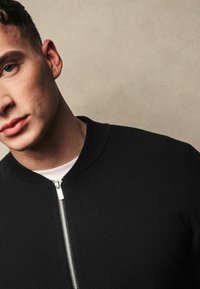 Černá bomber bunda s žebrovaným límcem, vybavená plným zipem a texturovanou pleteninou. Nositá přes bílou košili.