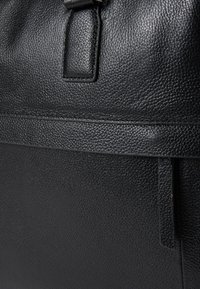 Crna kožna torba s teksturiranom površinom, koja ima mali, pravokutni ručni dio i vidljive šavove uz rubove.