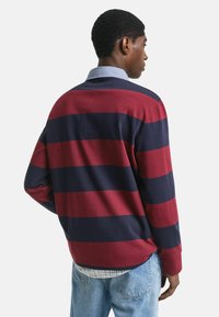 Mann in einem Rugbyshirt mit langen Ärmeln in breiten rot-blauen horizontalen Streifen, hellblauen Denim-Jeans und einem hellblauen Hemd mit Kragen darunter.
