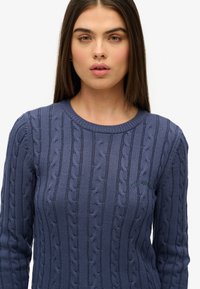 Navyblauw kabelgebreid sweater met een textuurpatroon, ronde halslijn en subtiele geborduurde logo op de borst. Lange mouwen.