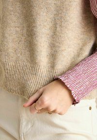 Beige Wollpullover mit geripptem Saum, über einem rosa gestreiften Hemd mit strukturierten Rüschen an den Bündchen getragen. Hand mit Ringen geschmückt.