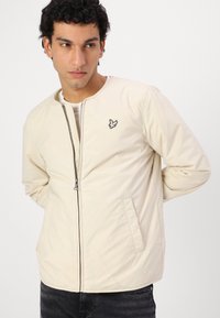 Lys beige jakke med rund hals og glidelås foran. Har sidelommer og en liten brodert logo på brystet. Glatt tekstur.