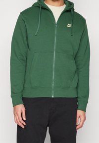 Grön zip-up huvtröja i mjukt tyg, med en känguruficka och en gul Nike-logga på vänster bröst. Huvan har snoddar.