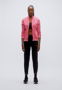 Ροζ αθλητικό μπουφάν με φερμουάρ και λευκές λεπτομέρειες, συνδυασμένο με μαύρο τοπ που φτάνει μέχρι τη μέση και μαύρο στενό παντελόνι. Τα μαύρα αθλητικά παπούτσια ολοκληρώνουν το σύνολο.