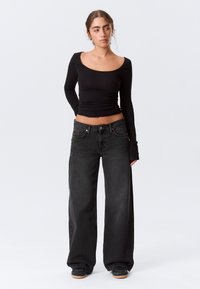 Jeune femme debout, portant un crop top noir à manches longues, un jean noir à jambes larges et des baskets noires avec des semelles couleur gomme.
