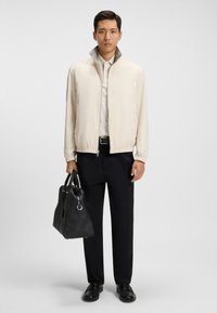 Jachetă crem deschis, cu fermoar și guler înalt, combinată cu un tricou polo alb, pantaloni negri și o geantă de mână texturată neagră. Textură netedă.