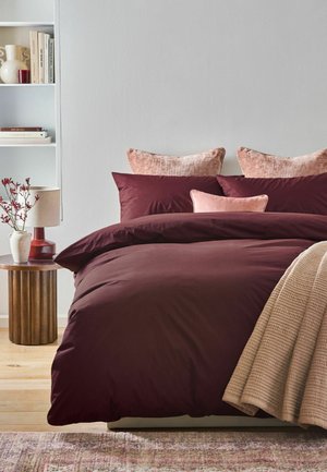 Bett mit dunkelroter Bettwäsche, pinken Kissen, beige Decke, Holznachttisch mit Lampe und Vase mit roten Blumen sowie weißem Bücherregal.
