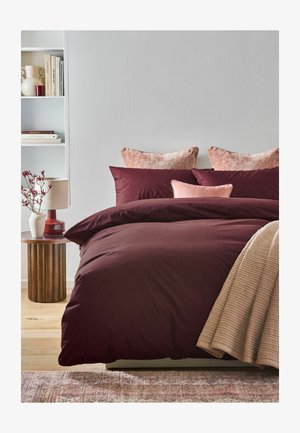 Bett mit dunkelroter Bettwäsche, pinken Kissen, beige Decke, Holznachttisch mit Lampe und Vase mit roten Blumen sowie weißem Bücherregal.