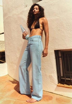 Žena s kovrčavom kosom koja nosi crni bikini top i svijetloplave traperice širokih nogavica, drži bocu pića i stoji bosa uz sunčan zid.