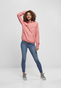 Giacca a vento rosa chiaro con cappuccio, dotata di una tasca frontale, polsini elasticizzati e orlo con cordino, abbinata a jeans blu aderenti e sneakers grigie.