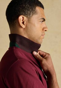 Burgundi polo särk kõrge kraega, millel on rohelised ja mustad triibud, tekstureeritud kangast ning lõdva lõikega, mudeli käsi reguleerimas kraed.