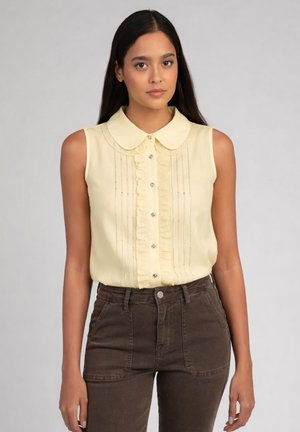 Jeune femme aux longs cheveux foncés, portant une blouse sans manches jaune pâle à volants et un pantalon marron taille haute, debout devant un fond uni.