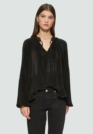 Blouse noire plissée avec un décolleté en V, un détail à nouer et des manches longues. Tissu texturé avec un ourlet froncé. Portée avec un pantalon noir ajusté.