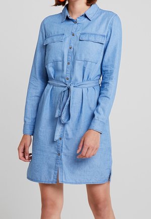 Vestido de mezclilla azul claro con mangas largas, cierre frontal de botones, bolsillos en el pecho y cinturón de lazo en la cintura; presenta un dobladillo curvado con aberturas laterales.