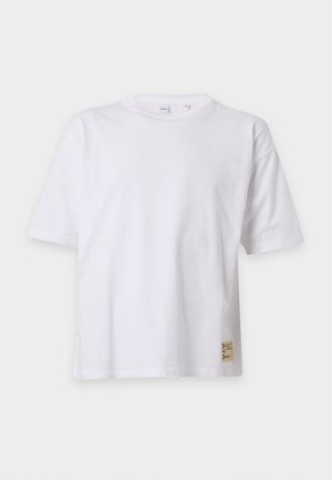 Homme debout, une main dans la poche, portant un t-shirt blanc uni, un jean bleu ample et des mocassins noirs et beiges sur fond blanc.