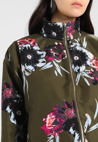Kwiatowa kurtka bomber w kolorze oliwkowym, z dużym czarnym i różowym wzorem kwiatowym. Wysoki kołnierz i zamek błyskawiczny z przodu. Gładka, błyszcząca tekstura.