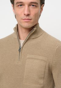 Beige strukturierter Strickpullover mit einem halben Reißverschluss-Kragen und einer Brusttasche, mit einem gerippten Muster und glatten Stoffdetails.